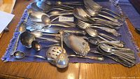 A variety of antique flatware and serving utensils laid out on a blue cloth including spoons, forks, ladles, and pie servers with detailed floral patterns.