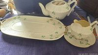 Photo showing the Belleek teapot, creamer, and serving plate together on a table. Visible shamrock decoration and gold trim present. Teapot lid missing, spout chipped.