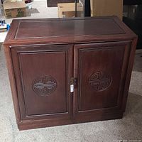 Front view of the closed dark rosewood cabinet with circular carved motifs on both doors, latch present, one handle missing.