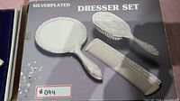 Silverplated dresser set components arranged on packaging showing hand mirror, hairbrush, comb, and additional brush/comb against a gray background with lot label.