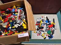 Box filled with assorted Lego pieces next to gray baseplate with scattered parts