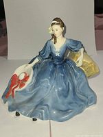 Front view of Royal Doulton Elyse figurine in blue dress holding white hat with red bow, seated on a pale yellow base.