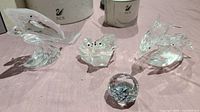 Four crystal figurines on light pink cloth: a pelican, a group of seals on a base, a dolphin, and a small faceted crystal ball with SCS etching.