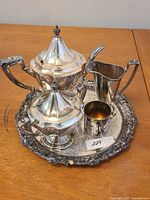 Silver tea set including teapot, sugar bowl with lid, creamer, small cup, and ornate tray on wooden table with lot number tag.