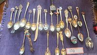 Full view of all spoons, fork, cutting utensil, and sugar tongs laid out on purple cloth.