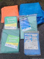 All tarps laid out on ground, including orange and blue used tarps, and green tarps in packaging.