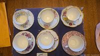 Top down view of all six teacups on matching saucers arranged on a blue placemat showing floral variety and gold trim
