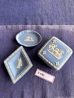 Three Wedgwood blue jasperware pieces displayed on purple fabric: square box with lid, diamond-shaped dish, oval dish. Fine white relief detail evident.