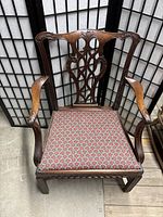 Front view of antique wooden armchair with floral seat upholstery showing carved wood detailing and armrests.
