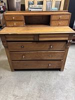 Front view of light brown wooden antique dresser showing 9 drawers including four small drawers on top, four larger drawers below, and an open middle compartment. Missing some drawer pulls and minor scratches visible.