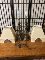 Pair of antique-style metal table lamps with fabric shades featuring bead trim, placed on wooden surface with a folding screen background.