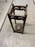 Front and top view of dark brown wooden umbrella stand with ornate carved panels and four turned finials at corners. Visible metal drip tray base shows patina and wear.