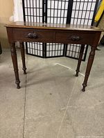 Full view of the antique wooden table showing rectangular top, two drawers with carved handles and turned legs.