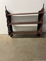 Front view of dark brown wooden wall-mounted shelf with three shelves and intricate side carvings.