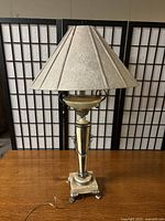 Full view of lamp unlit, showing fabric shade, gold-tone metal body with decorative chains, and claw footed base on wooden surface with black folding screen background.