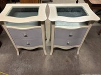 Pair of white nightstands side by side on concrete floor, showing glass tops and two drawers each with curved fronts and silver pull handles.