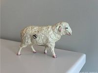 Front left angled view of the antique cast iron sheep coin bank showing white paint, pink hooves and ears, and blue eyes with some wear spots.