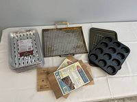 All items displayed on a white surface: cupcake pan, cooling rack, perforated grilling sheet with wooden handle, multiple other metal items, and three cedar grilling planks with packaging.