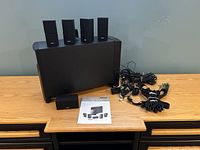 Front view of powered subwoofer with four cube speakers on top, center speaker in front, instruction manual and bundled cables laid out on table.