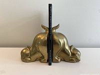 Pair of heavy brass cat bookends holding the book 'Martin Leman's Painted Cats'. Book spine visible with title and publisher Pelham Books.