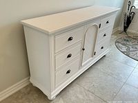 Angled view of full dresser showing white finish, six drawers, central cupboard door and black handles.