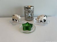 Two ceramic pig shakers on clear flower-shaped dishes, silver metal candle holder, and green glass star-shaped wax warmer on clear round base.
