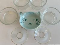 Four clear glass votive holders, one ceramic cat face dish, and two gold-rimmed glass bobeches arranged on a white surface.