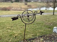 Side view of the metal armillary sphere garden stake placed outdoors in the grass with a pond in the background.