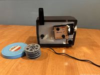 Kodak Brownie 8 Model 10 projector powered on with lamp lit, two 8mm film reels and blue plastic case displayed on wooden table.