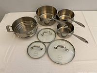 Photo of three stainless steel saucepans of different sizes with three corresponding glass lids and a stainless steel steamer basket.