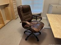 Brown leather swivel office chair with visible wear and cracks on seat, armrests, and headrest, shown at an angle in office environment.