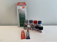 Photo showing 6 green-handled paintbrushes in original packaging, 10 small jars of Tamiya acrylic paints in various colors, and 2 tubes of plastic model cement (one red and one blue).