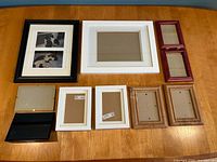 Nine picture frames arranged on a wooden table showing variety in sizes and colors including black, white, wood, and burgundy.