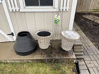 Photo showing two white wicker planters with liners on black metal plant stands, a large black ribbed plastic planter, black garden edging panels stacked beside them, and a coil of tomato wire with packaging attached.