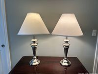 Pair of Almira brushed stainless steel table lamps on a wooden surface, showing the full lamps with white conical fabric shades.