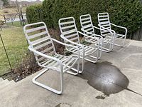 Four white metal patio chairs shown outdoors on a concrete patio, arranged in a row, highlighting the cantilevered tubular metal frame and horizontal slats.