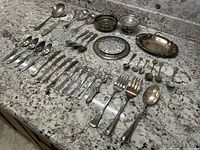 Overview of the full selection of silver flatware and serving ware laid out on a granite kitchen counter.