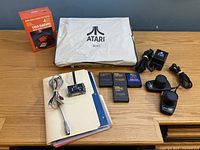 Wide view of all lot contents on wood surface: boxed Star Raiders, dust cover, power adapter, cables, joysticks, manuals, cartridges