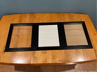Three large black photo wall frames placed side by side on a wooden table, showing front view with glass and backing board visible.