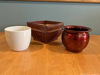 Three planters displayed from front angle on wooden surface: red ceramic square pot with white detailing, shiny red metal rounded pot, white ceramic eggshell textured pot.