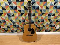 Full view of the Vintage Raven acoustic guitar standing against a colorful geometric patterned background, showing the natural wood finish and dreadnought body shape.