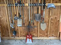 Ten long-handled garden tools and a grey plastic hose holder hung on wooden wall