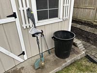 View of the Yard Works string trimmer, hand pruner leaning against a beige shed wall, and black garbage pail on concrete