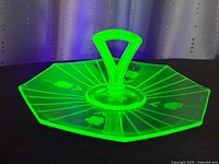 Front angle of green uranium glass dessert tray showing octagonal shape, central handle, and floral wheel cut pattern glowing under black light.