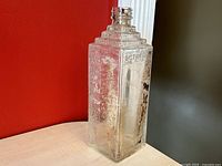 Full view of the clear glass embalming bottle, showing its square shape and stepped neck design with some residue inside.