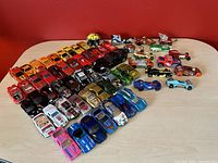 Photo shows all 60 assorted Hot Wheels cars arranged on a light wooden surface against a red wall. Cars are grouped by color and type including reds, yellows, blues, greens, pinks, blacks, and multi-colored specials.
