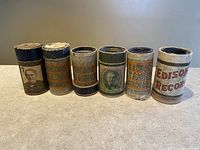 Front view of 6 antique Edison Blue Amberol Records cylinders in their original cylindrical cardboard containers, showing various label designs and coloration.