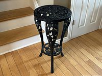 Front angle view showing intricate grape vine and leaf designs on the round top, legs, and lower shelf of the black cast iron plant stand. The removable table top is visible.
