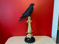 Full view of gold and black resin raven bird statue with ornate gold column and black skull base against red background.