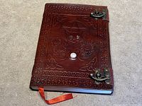 Front cover of brown embossed leather journal showing pentagram and cat design with jewelled eyes, bronze metal clasps, and orange bookmark ribbon partially visible.